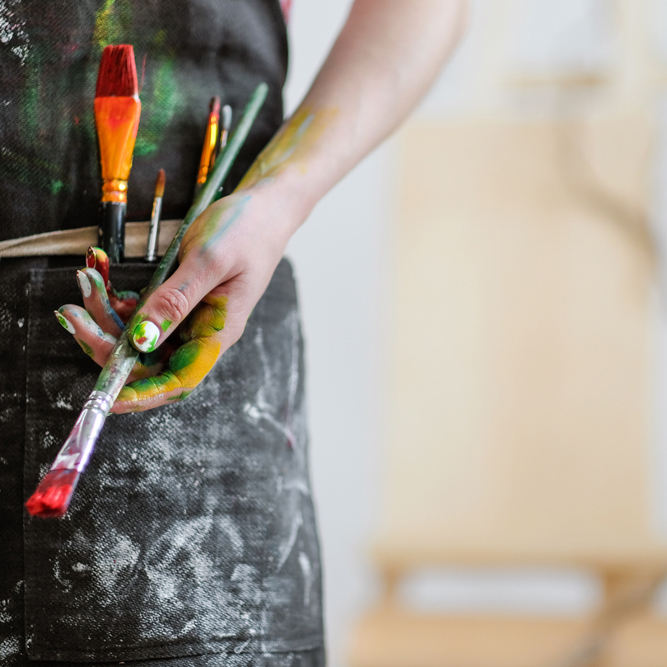 Mains d'une artiste peintre tenant ses pinceaux, tablier taché de couleurs